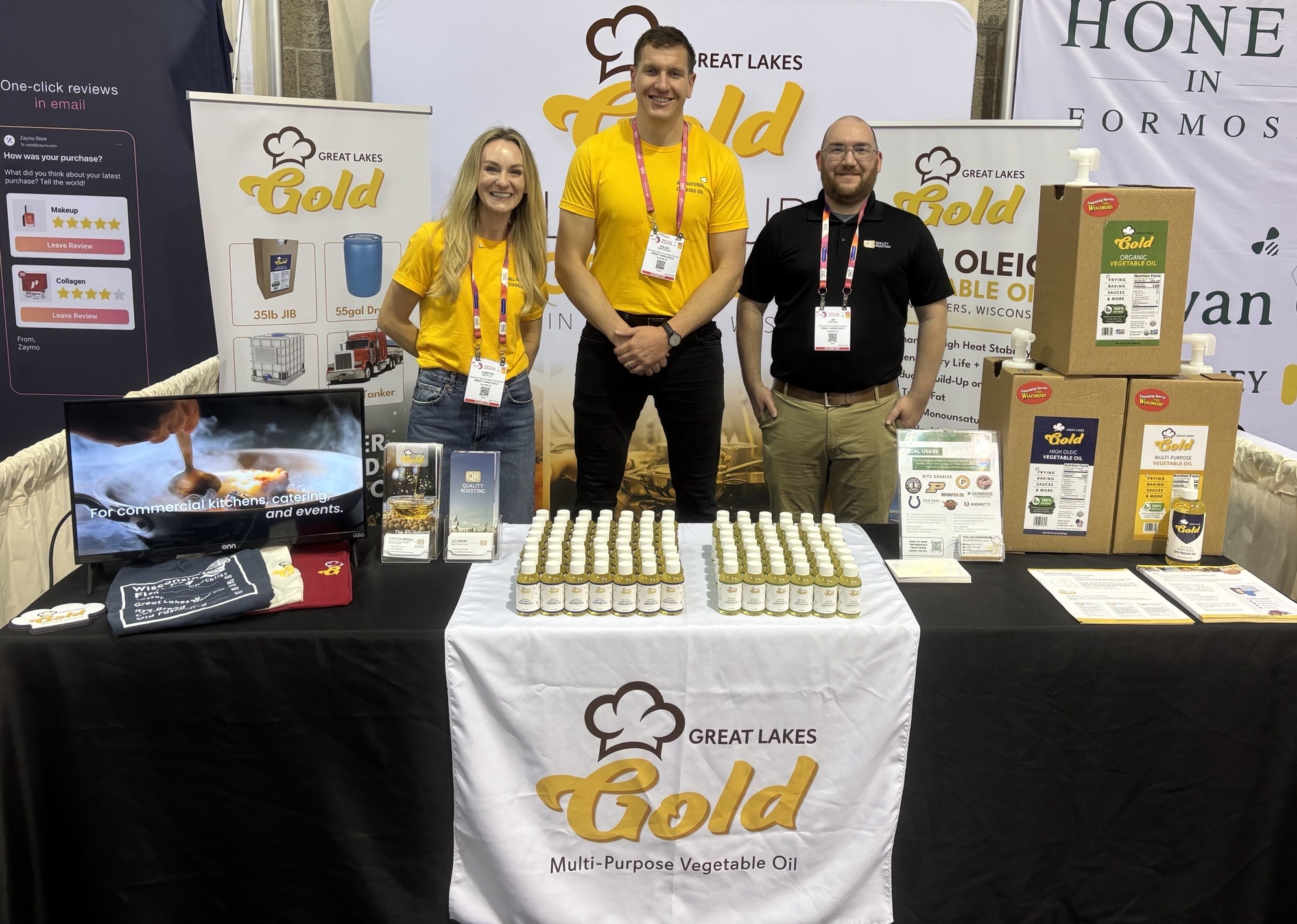 Three booth staff in yellow and black shirts stand behind a Great Lakes Gold display at a trade show booth, with bottles lined on the table and stacked boxes to the side.